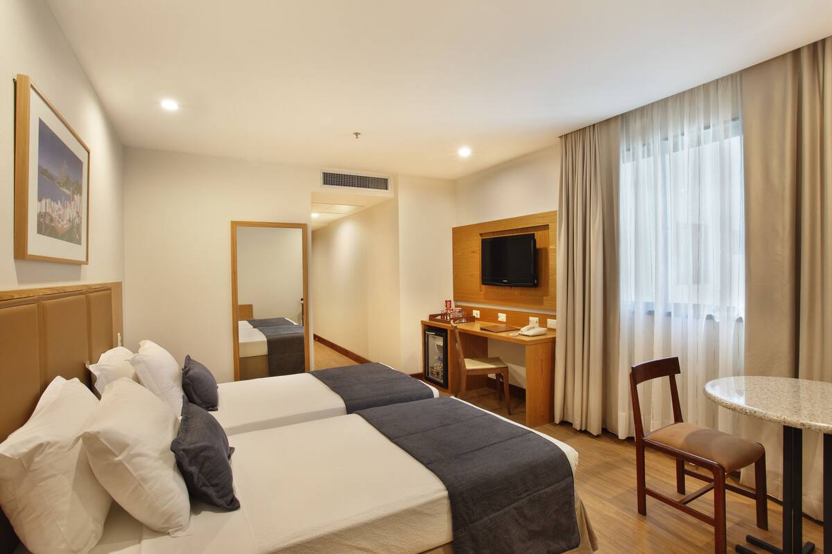 A hotel room with two beds, a desk, a chair, a TV, and a window with curtains.