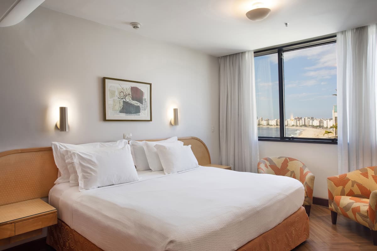A hotel room with a large bed, a chair, a window with a city view, and a framed picture on the wall.