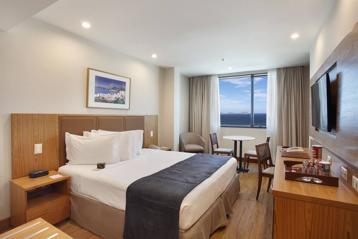 A hotel room with a large bed, a desk with a flat-screen TV, a chair, a small table, and a window with a view.
