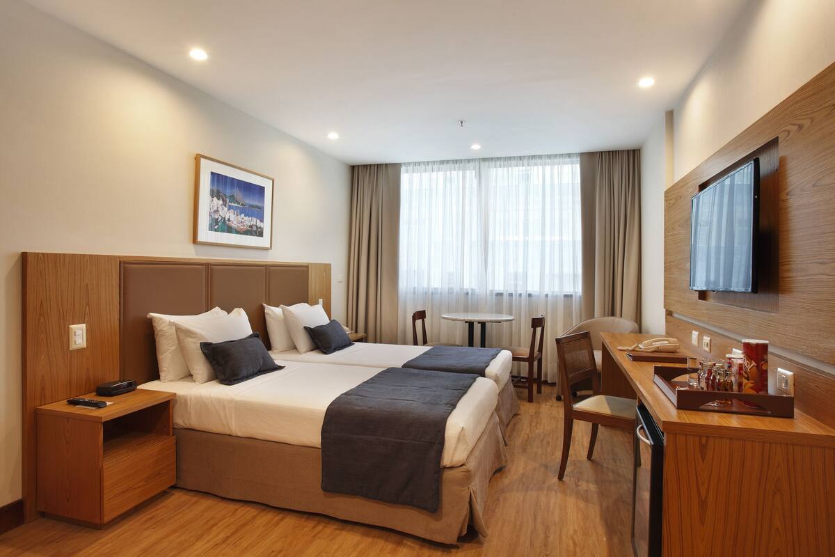 A hotel room with two beds, a desk, a chair, a TV, and a window with curtains.