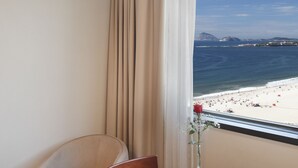 Una habitación con una mesa redonda, una silla, un jarrón con una flor y una ventana que ofrece vistas a una playa y montañas, además de una cortina.
