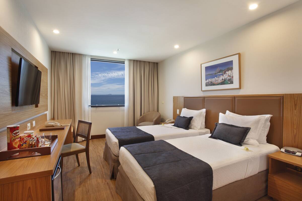 A hotel room with two beds, a desk with a TV, a chair, and a window with a view of the sea.