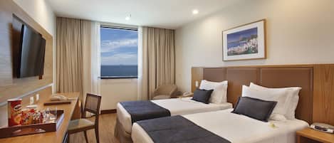 A hotel room with two beds, a desk with a TV, a chair, and a window with a view of the sea.