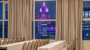 Un salón de conferencias con mesas y sillas, ventanas grandes con cortinas y vistas a un paisaje urbano nocturno.