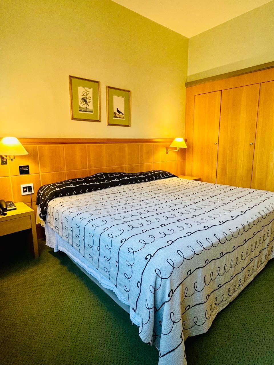 A hotel room with a bed, two bedside lamps, a nightstand, a wooden wardrobe, and two framed pictures on the wall.