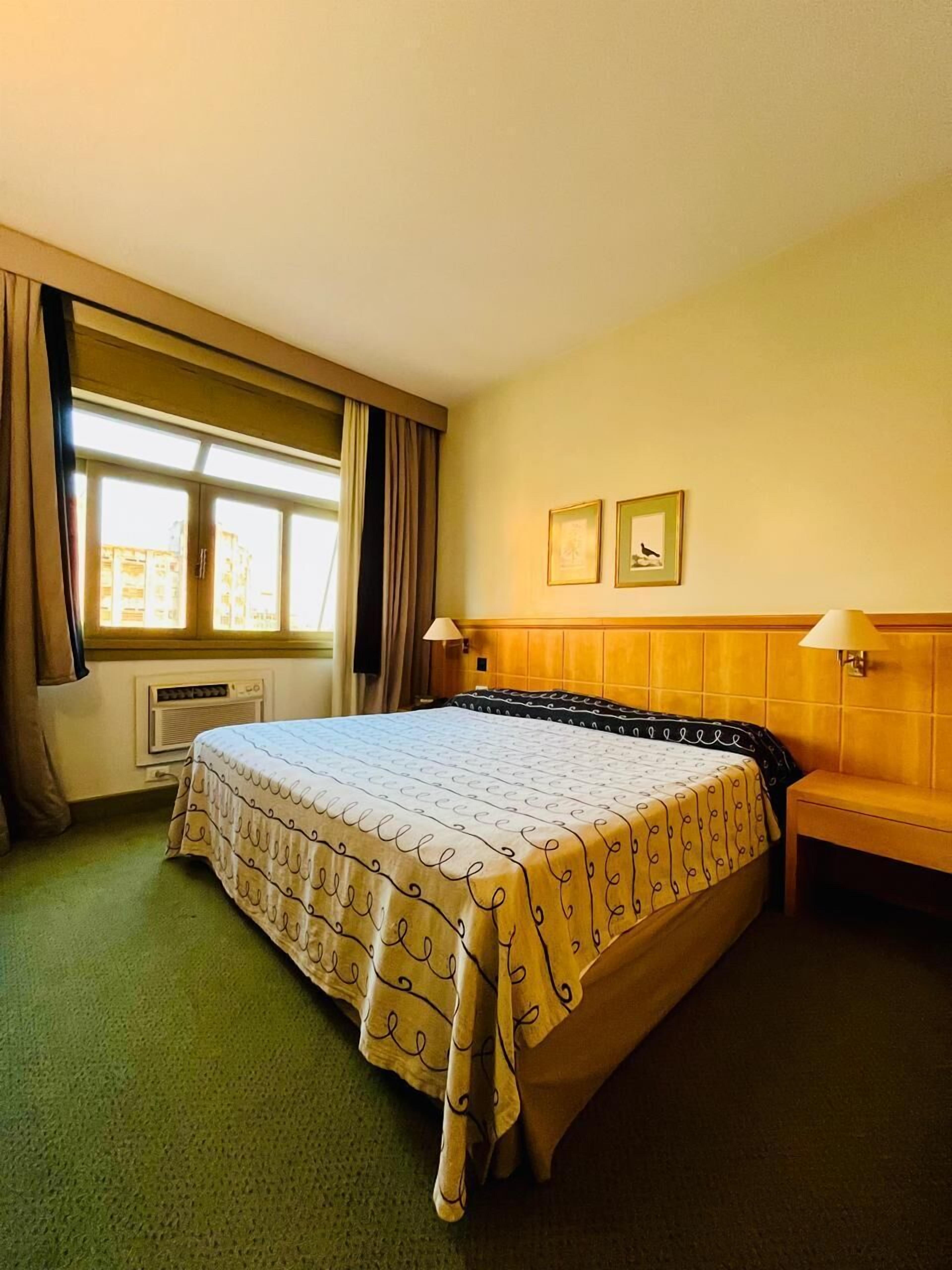 Een hotelkamer met een groot bed, een raam met gordijnen, een radiator, twee wandlampen en twee ingelijste foto's aan de muur.