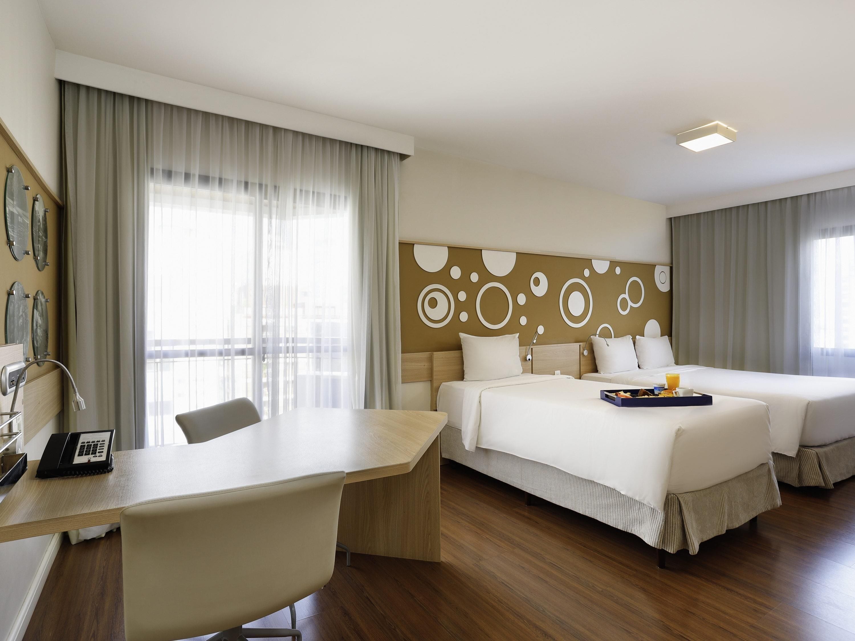 Ein Hotelzimmer mit Schreibtisch, zwei Betten, einer Wand mit kreisförmigen Mustern und einem Fenster mit Vorhängen.