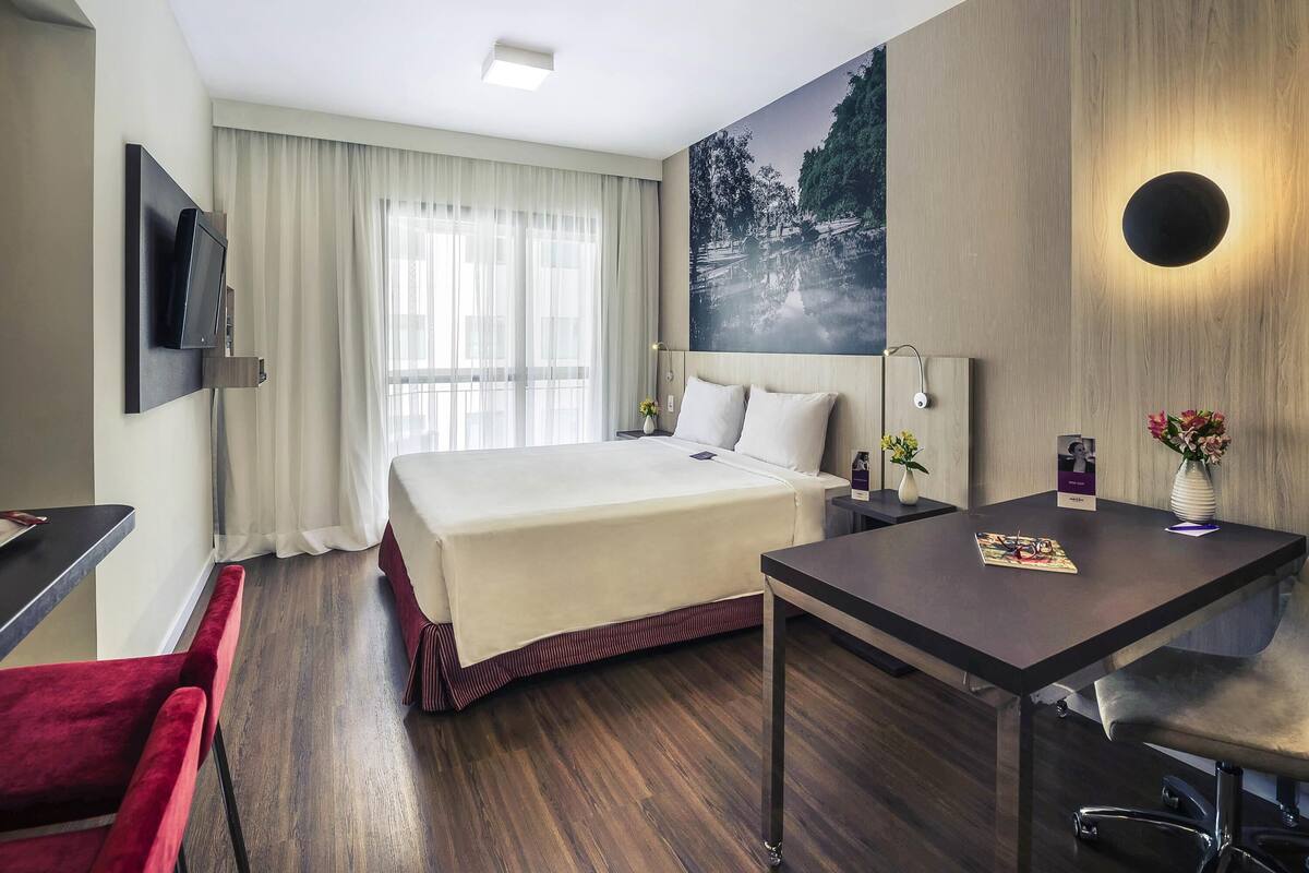 Une chambre d’hôtel avec un lit, un bureau, une chaise, un téléviseur fixé au mur, un tableau au mur et une fenêtre avec des rideaux.