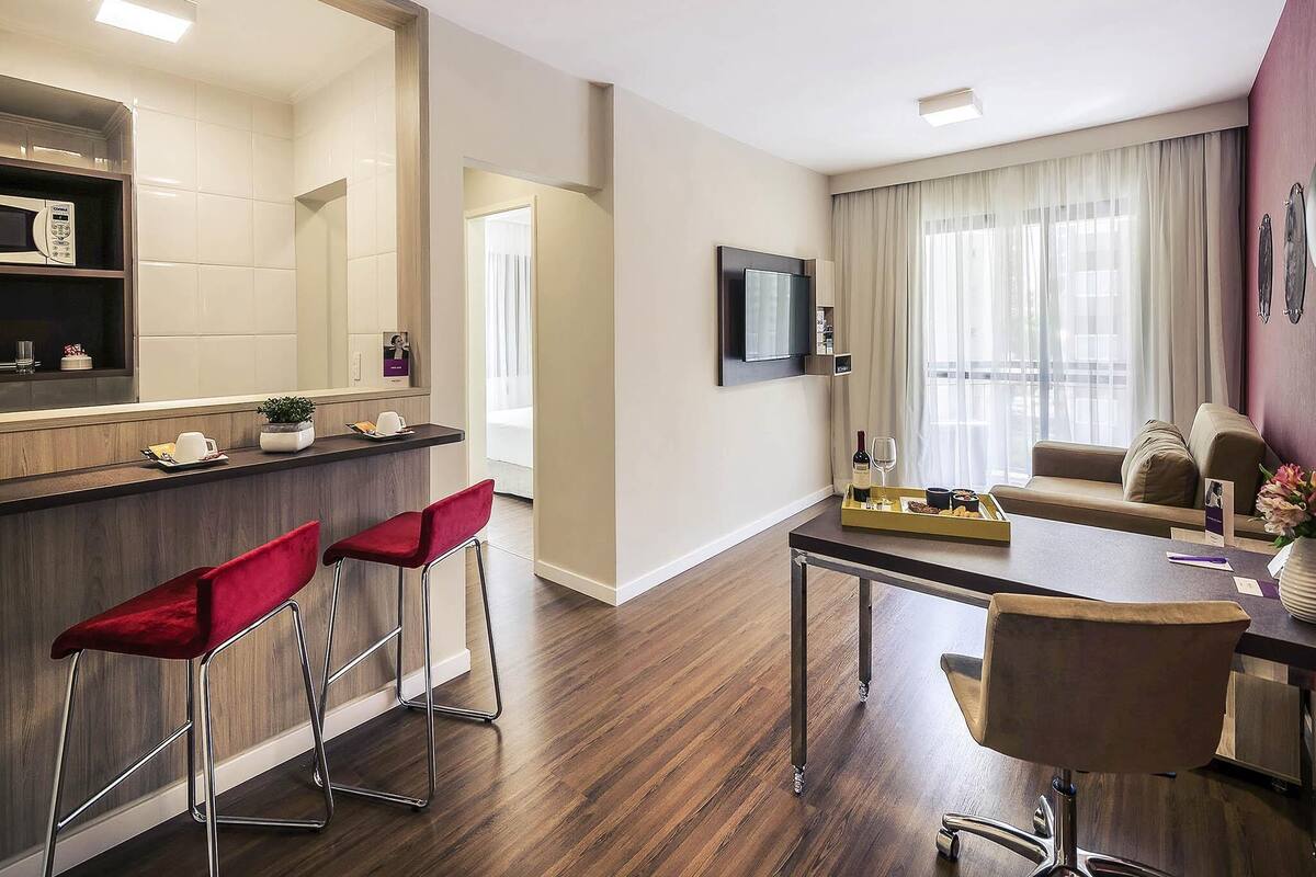 Une chambre d’hôtel moderne avec un plancher en bois, un coin repas avec une table et des chaises, un bar avec des tabourets, un téléviseur fixé au mur et une grande fenêtre avec des rideaux.