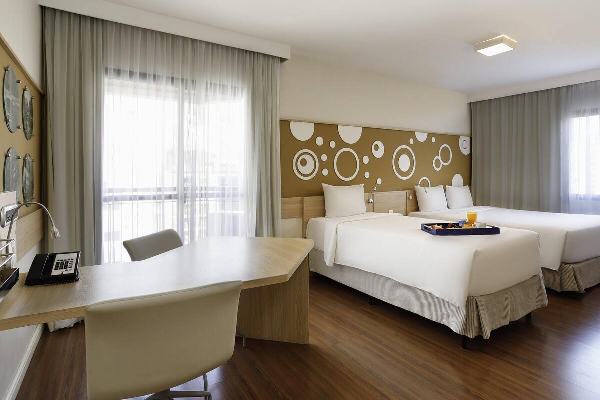 Une chambre d’hôtel avec deux lits, un bureau et une grande fenêtre avec des rideaux.