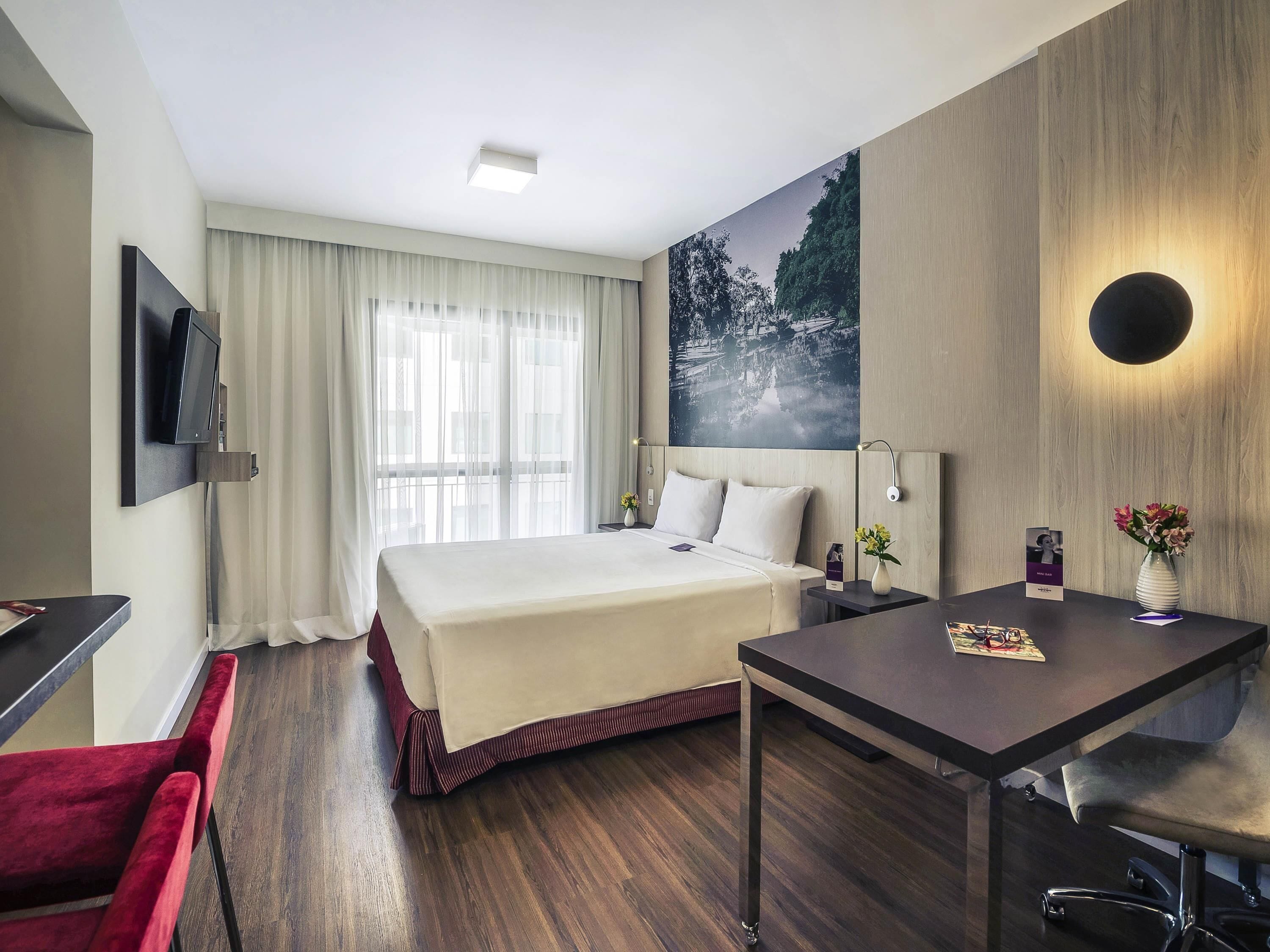 Ein Hotelzimmer mit Bett, Schreibtisch, Sessel, Wandfernseher, Vorhängen an einem Fenster, einem Bild an der Wand und einem Nachttisch mit Blumen.'