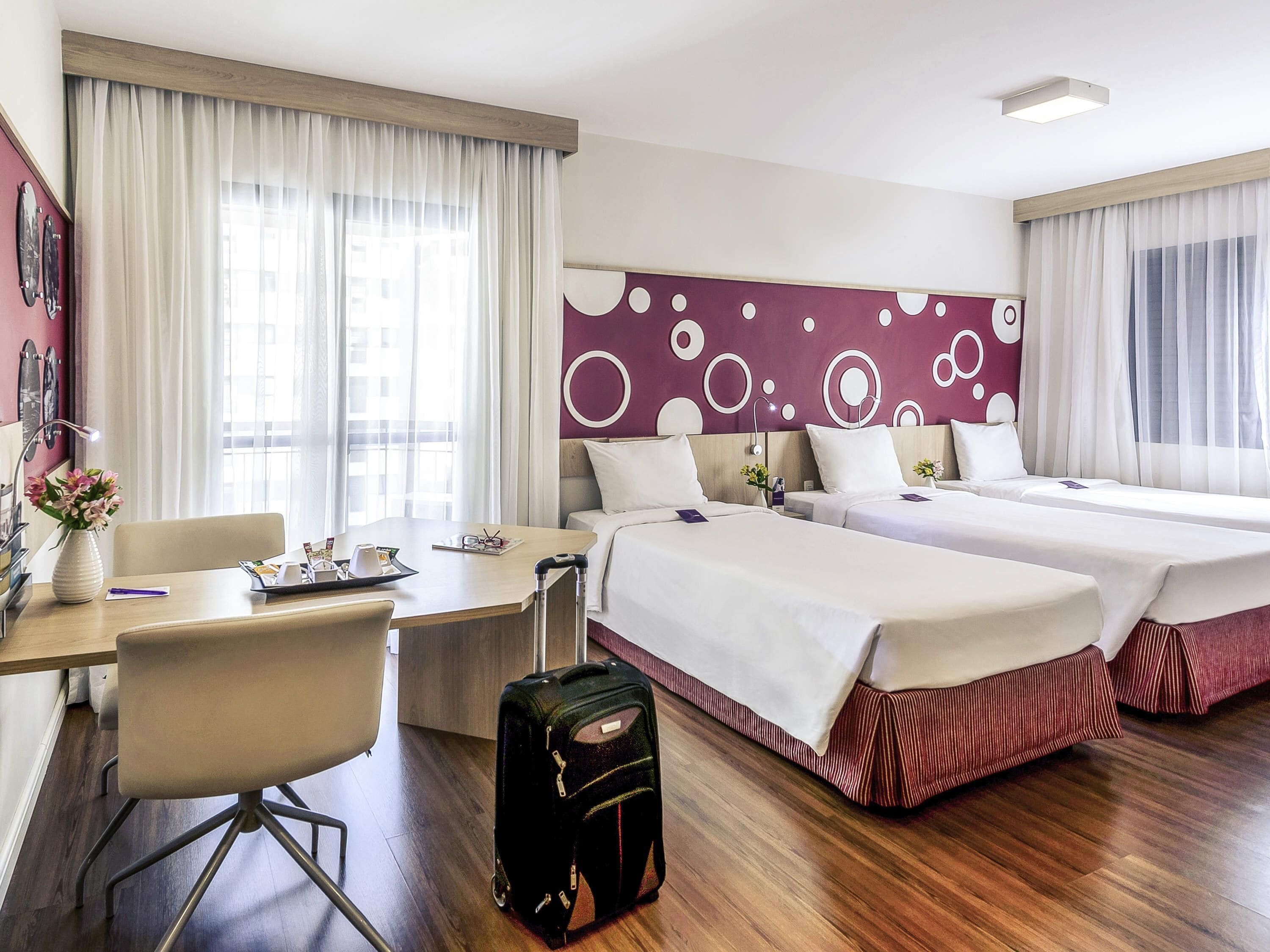 Quarto de hotel com duas camas, uma escrivaninha, uma cadeira e uma mala.