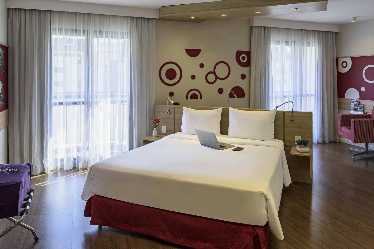 Une chambre d’hôtel avec un grand lit, un bureau, une chaise et une fenêtre avec des rideaux légers.