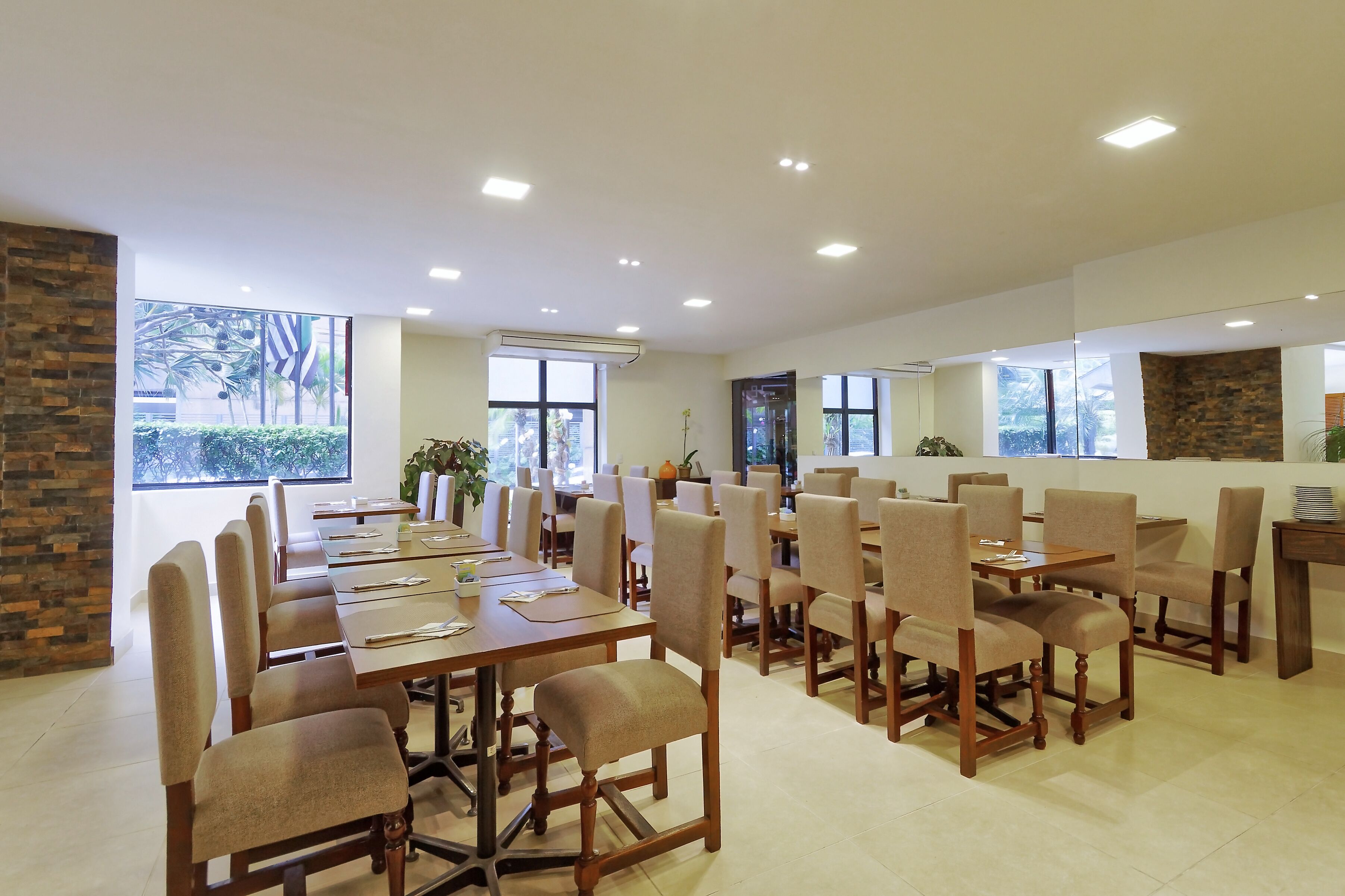 A spacious dining area with multiple tables and chairs, large windows with a view of greenery, and a modern ceiling design.
