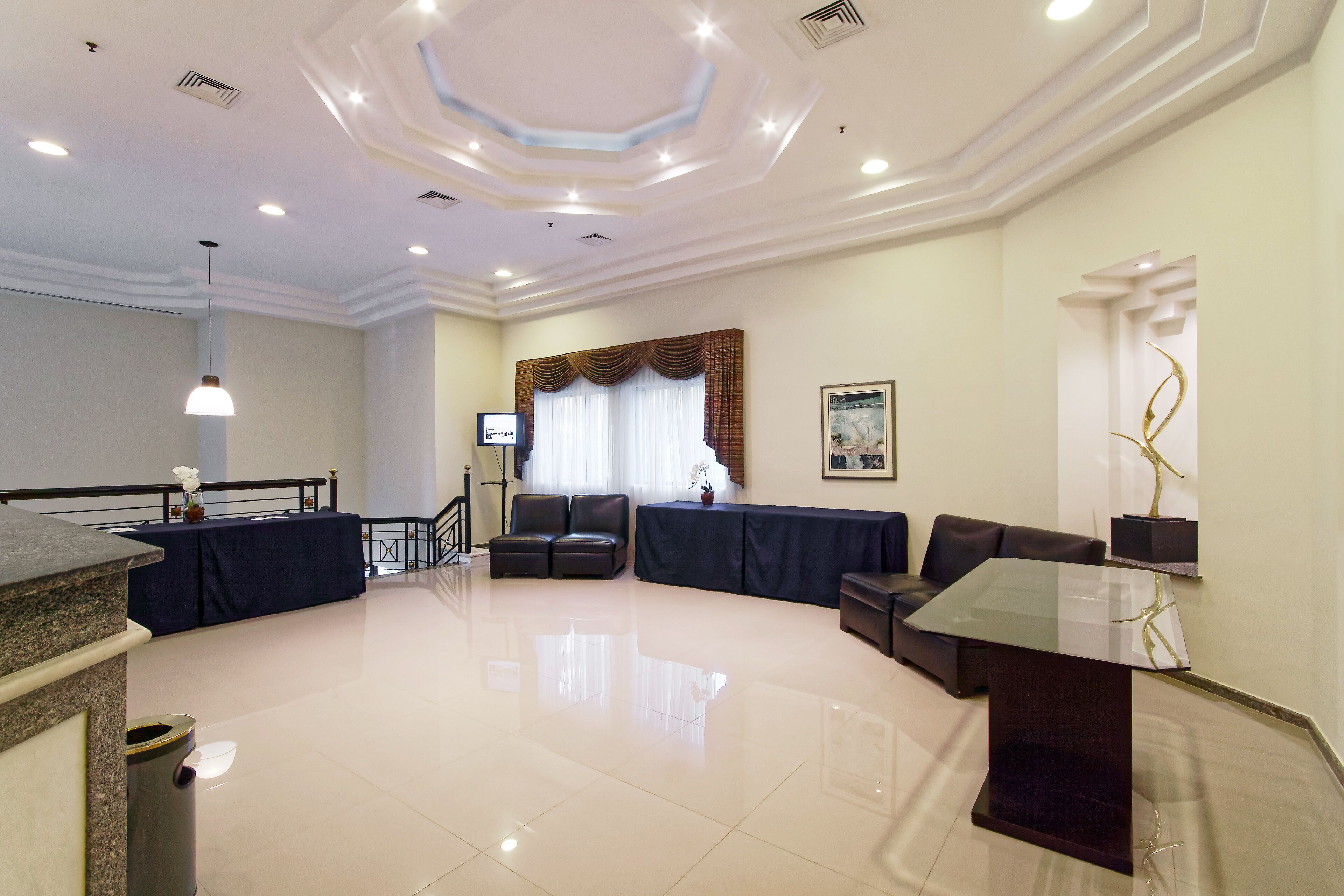 A spacious hotel lobby with a modern design, featuring a seating area, a reception desk, and a large window with curtains.