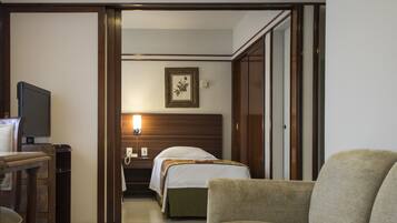 Quarto de hotel com cama, uma cadeira, escrivaninha com televisão e mesa de cabeceira com abajur.
