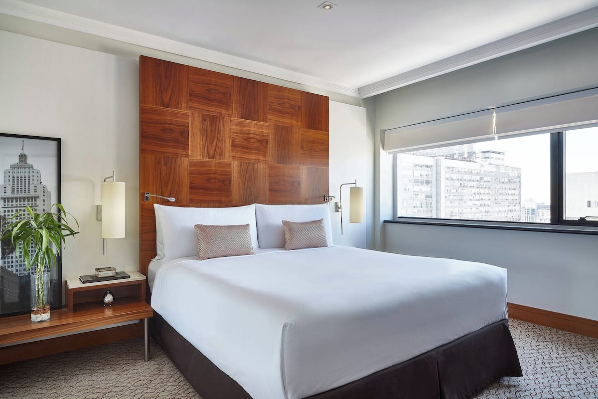 A modern hotel room with a large bed, a wooden headboard, a city view, and a nightstand with a lamp and a plant.