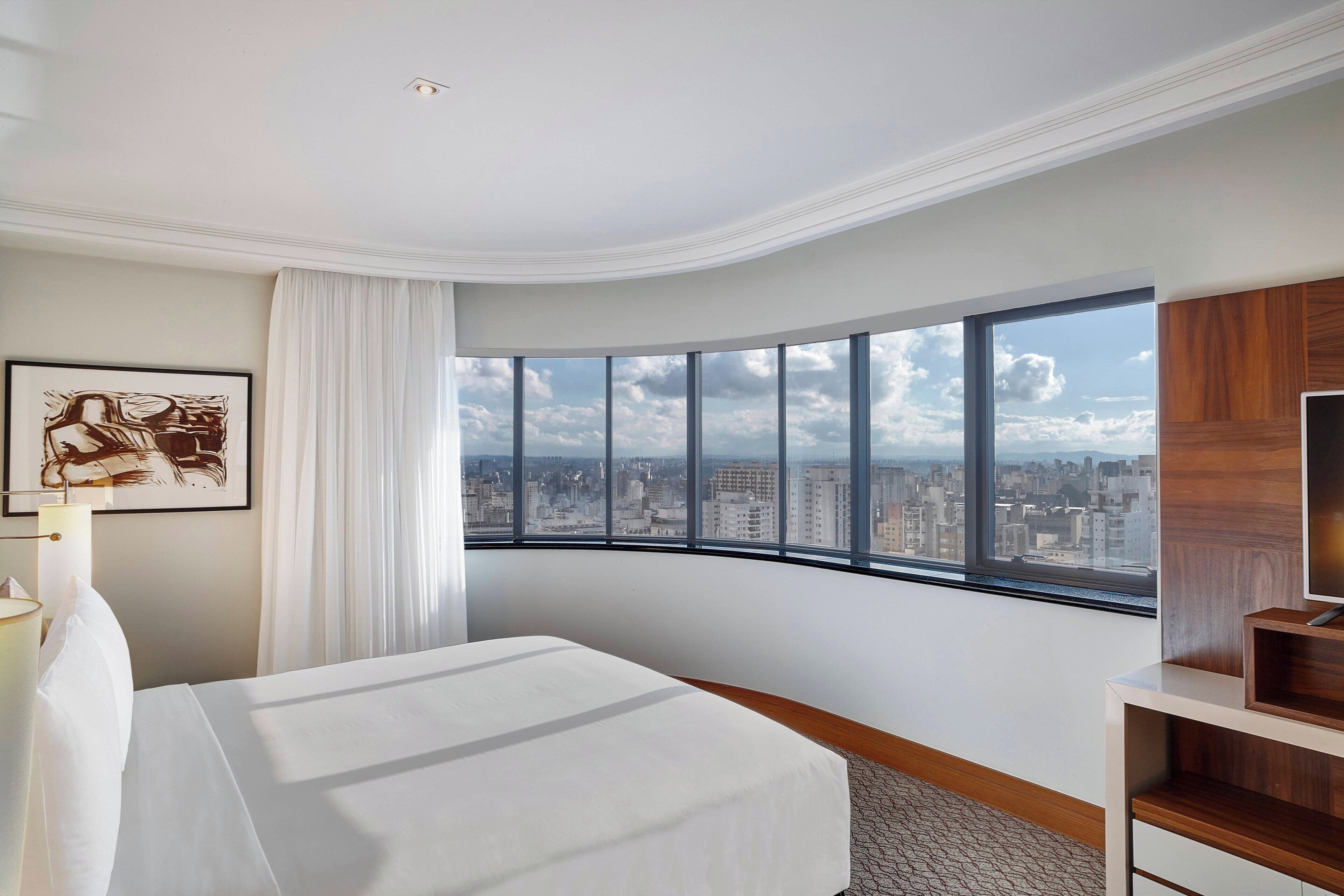 A hotel room with a curved window offering a city view, a neatly made bed, a wooden sideboard, and a framed artwork on the wall.
