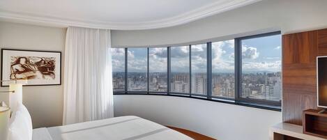 A hotel room with a curved window offering a city view, a neatly made bed, a wooden sideboard, and a framed artwork on the wall.