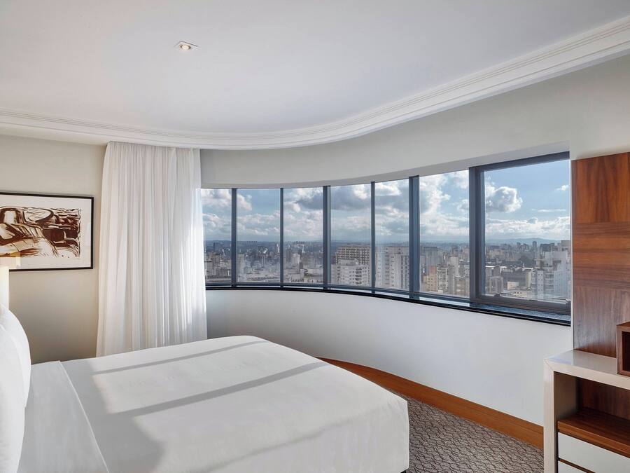 A hotel room with a curved window offering a city view, a neatly made bed, a wooden sideboard, and a framed artwork on the wall.