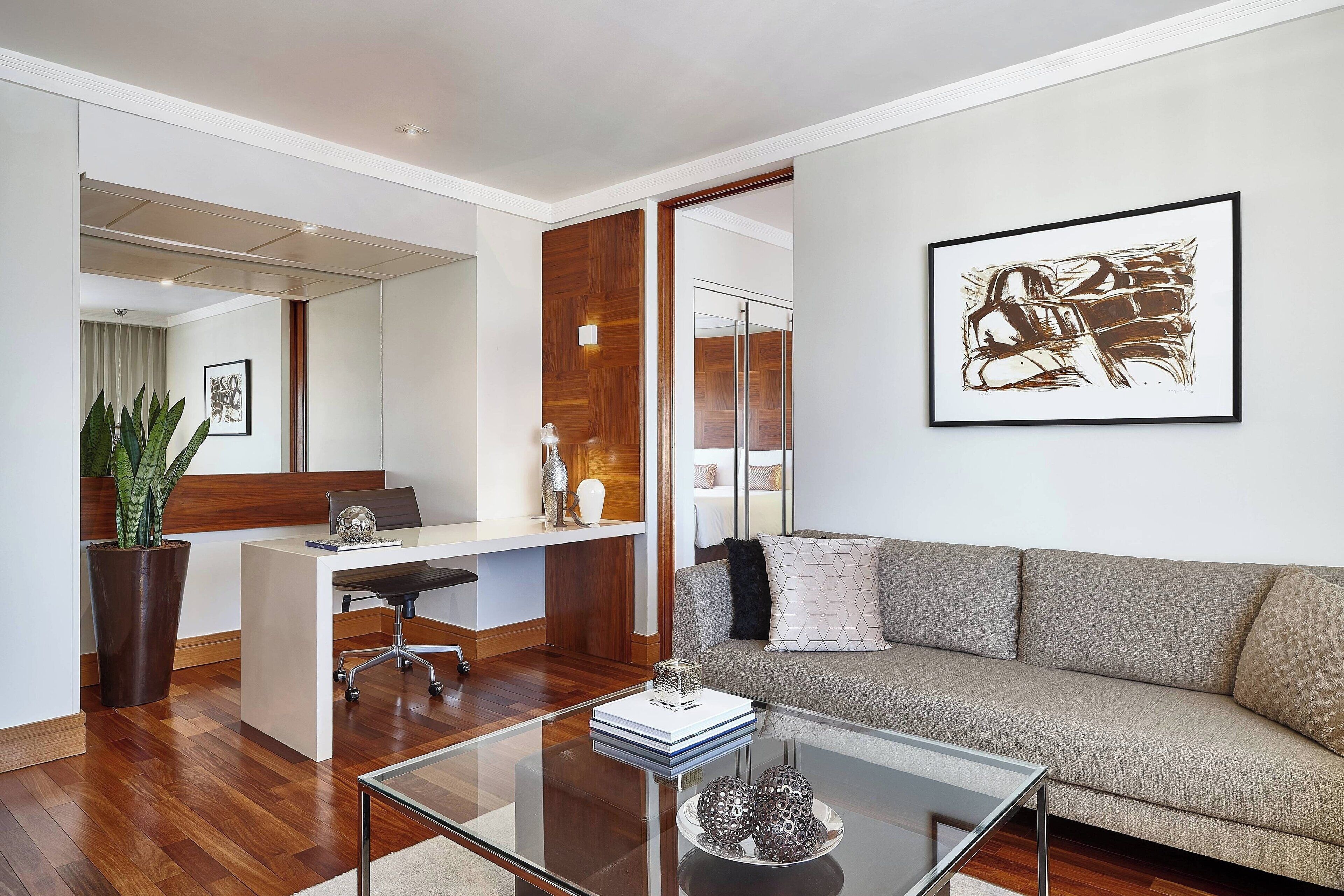 A modern living room with a glass coffee table, a sofa, a desk with a chair, and a large abstract painting on the wall.