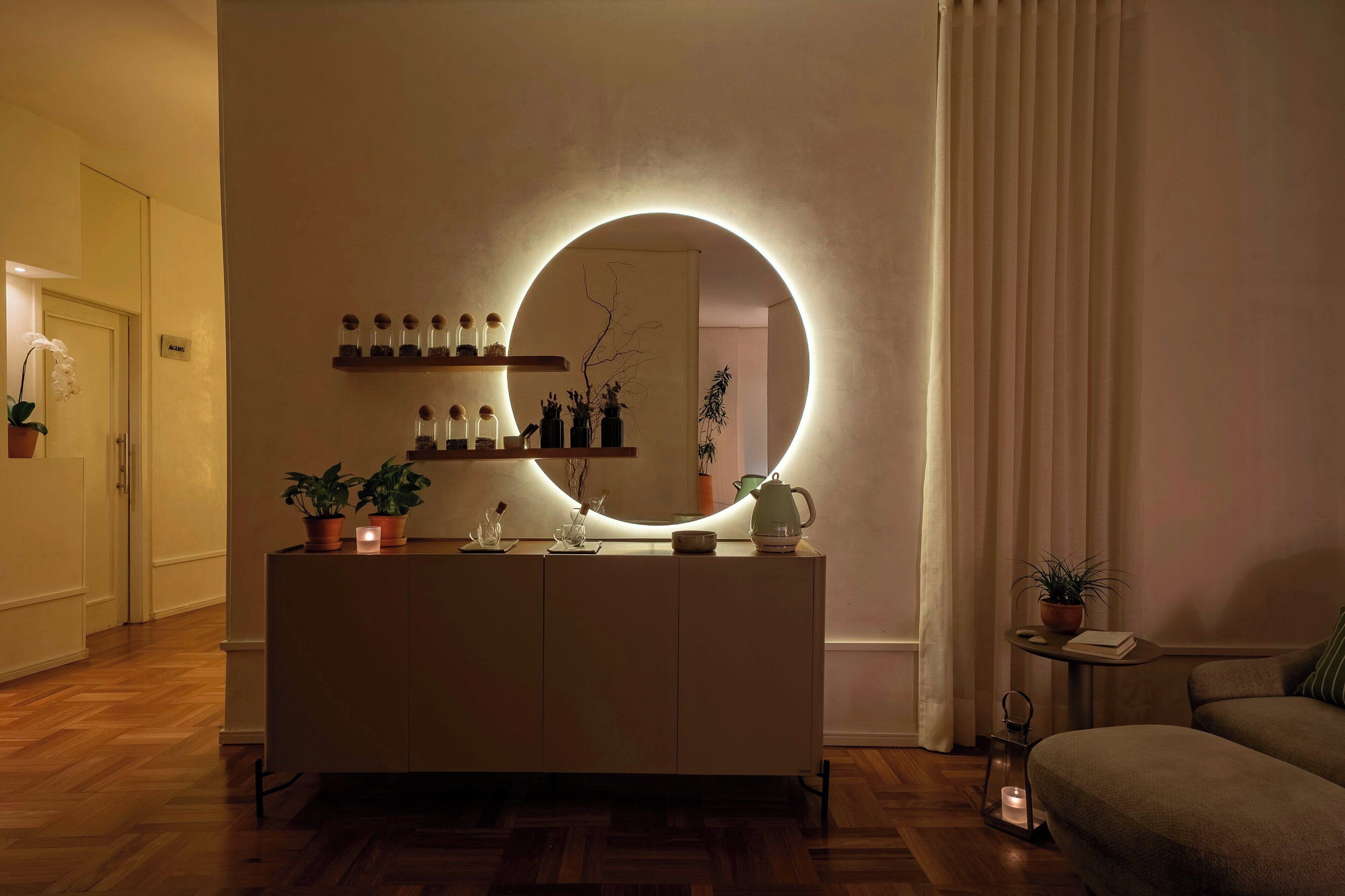 Quarto moderno com piso de madeira, um console central com objetos decorativos, um espelho redondo com iluminação e uma pequena mesa com uma planta.