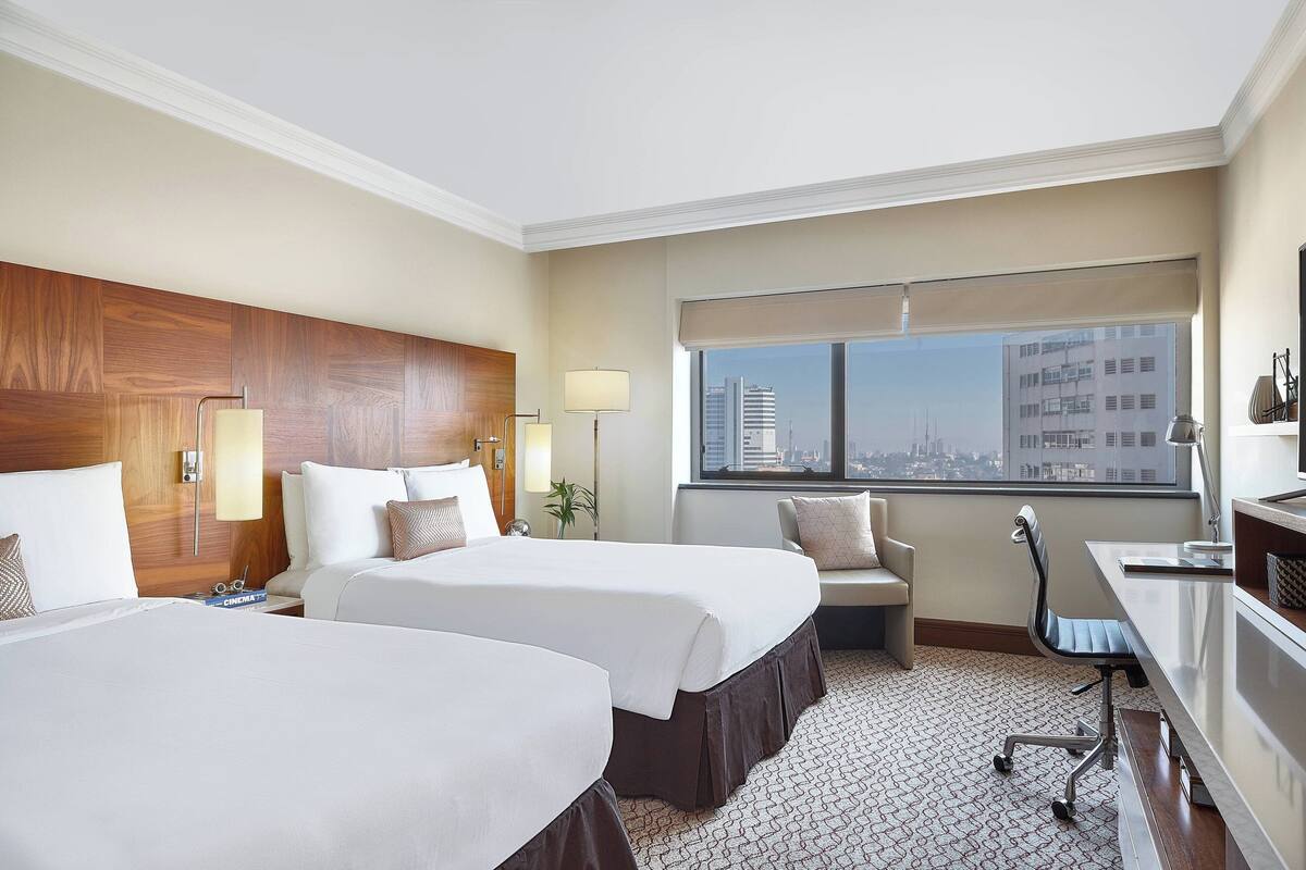 A hotel room with two beds, a desk, a chair, a lamp, and a large window with a city view.