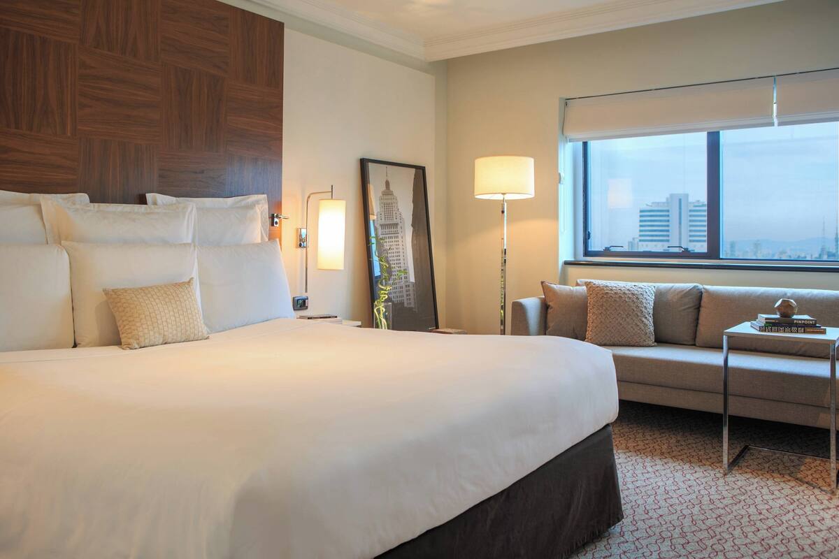 A hotel room with a large bed, a sofa, a standing lamp, a framed picture, and a window with a city view.