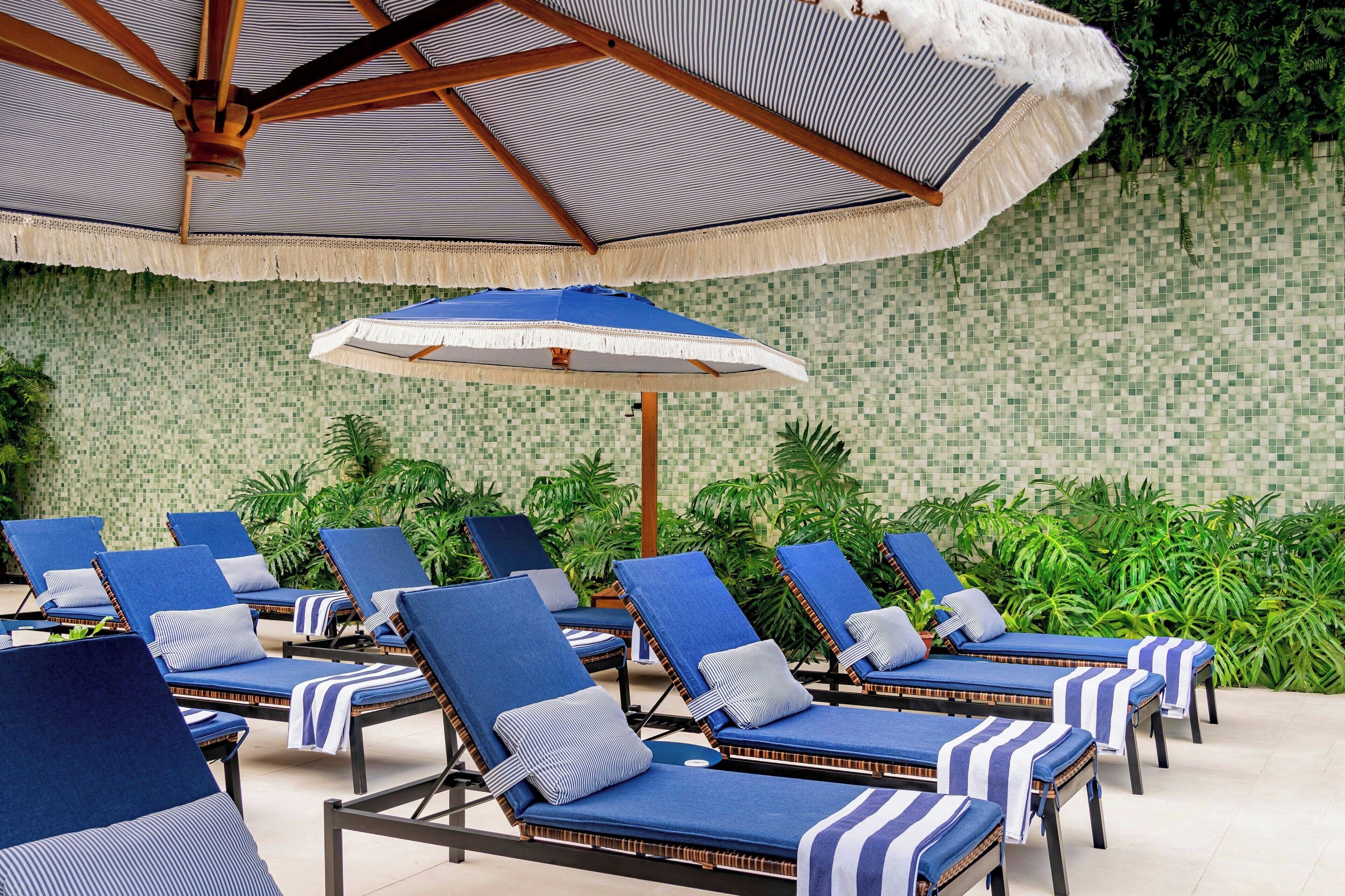 A poolside area with lounge chairs, a large umbrella, and a tiled wall with greenery.