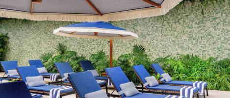 A poolside area with lounge chairs, a large umbrella, and a tiled wall with greenery.