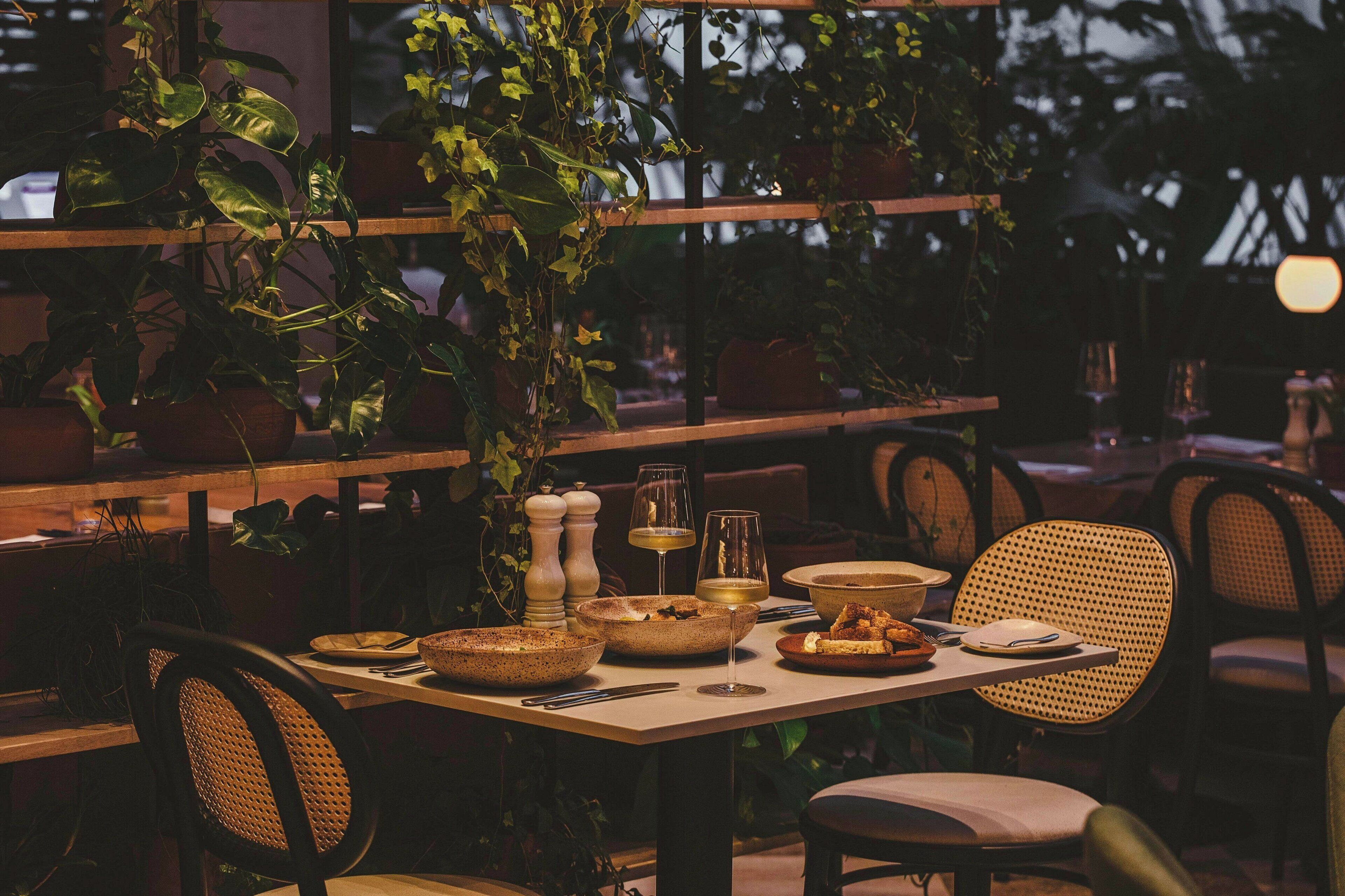 Comedor con una mesa dispuesta para dos personas, rodeada de sillas de mimbre, plantas en macetas y un ambiente cálido.