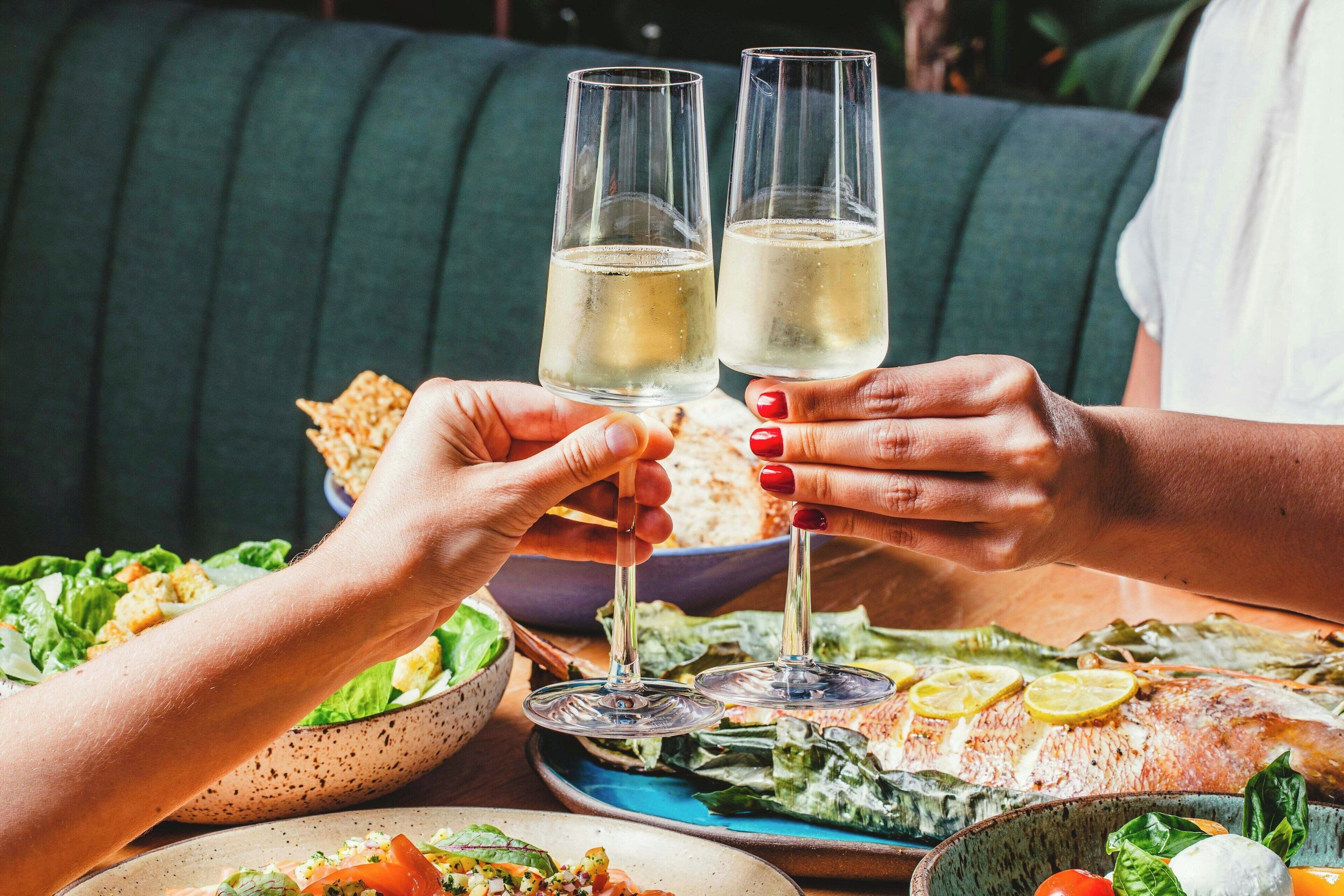 A dining table with a fish, salad, and bread. Two people are toasting with wine glasses.