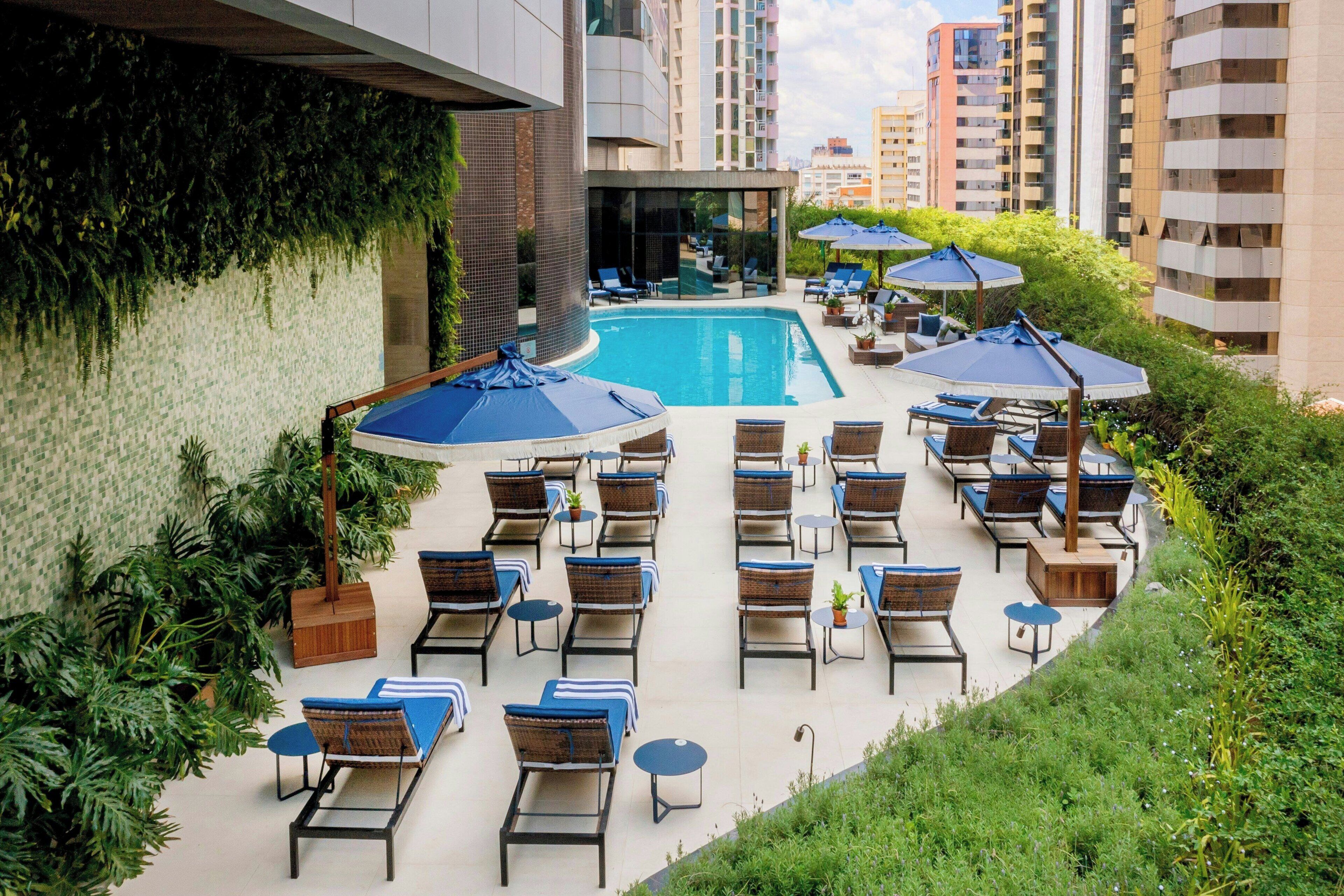 A rooftop pool area with lounge chairs and umbrellas, surrounded by greenery and high-rise buildings.