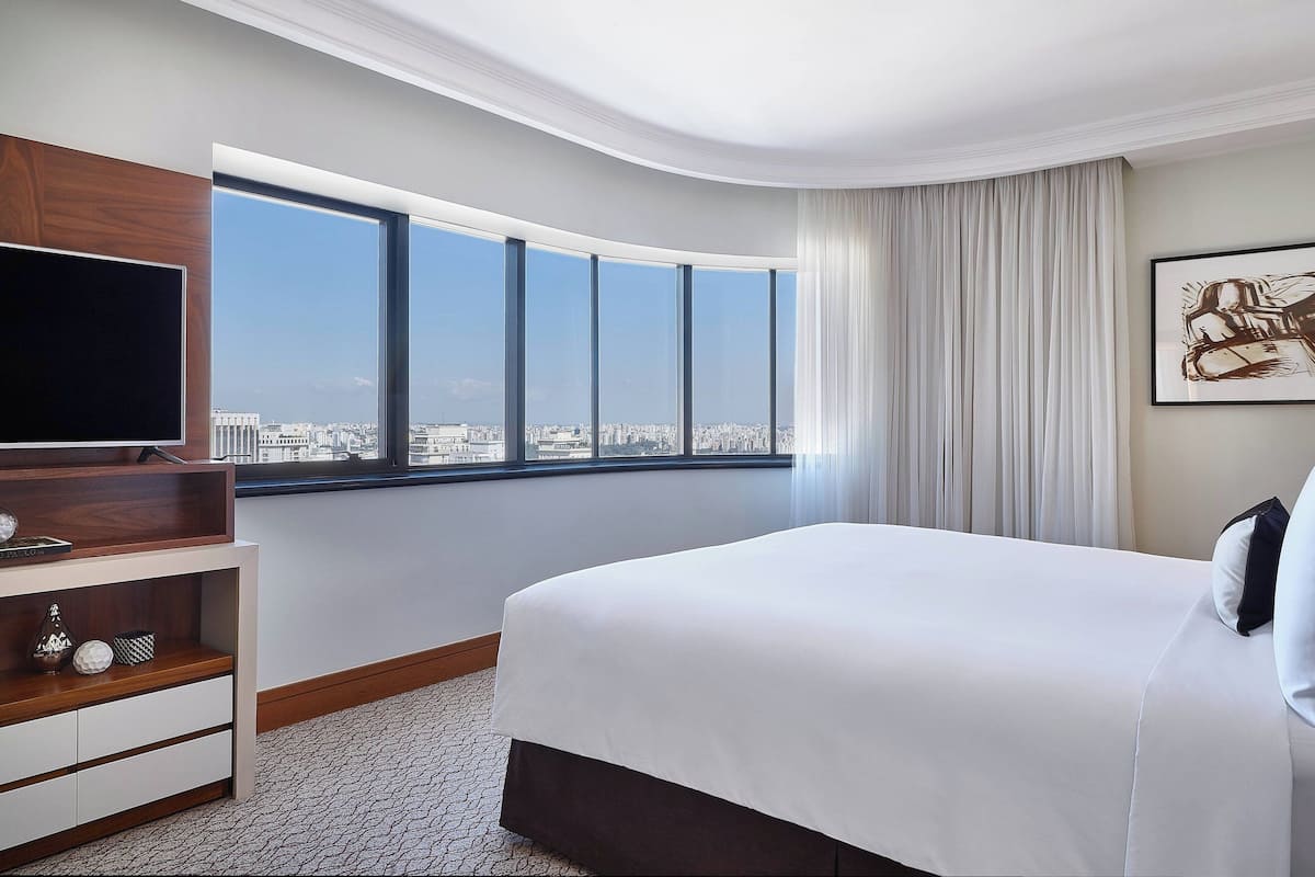 A hotel room with a large bed, a TV on a wooden stand, a city view, and a large window with curtains.