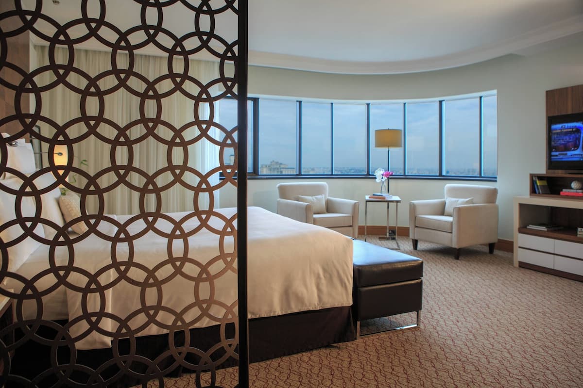 A hotel room with a large bed, a seating area with two chairs, a TV, and a view of the city through large windows.