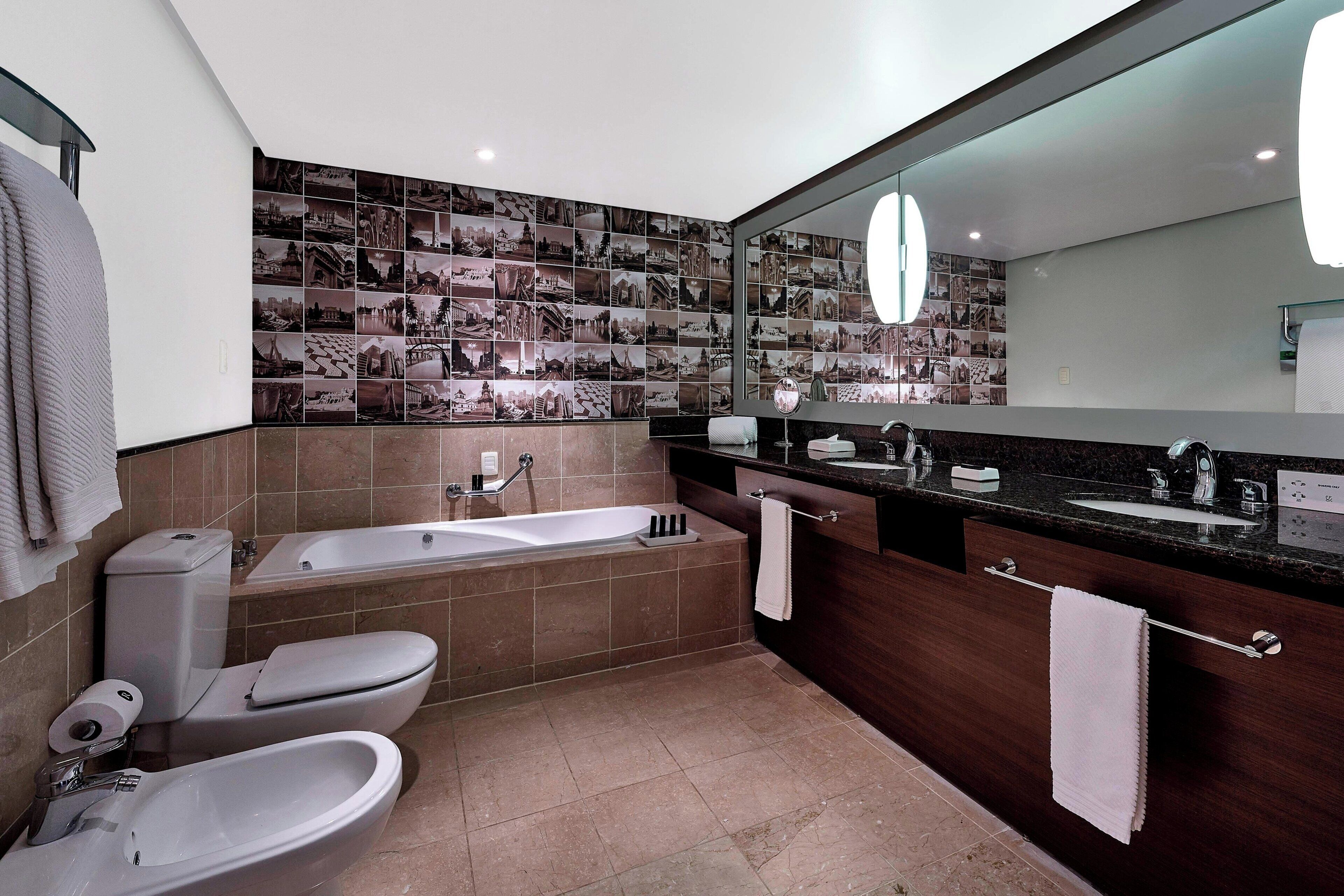 Un baño moderno con bañera, inodoro y bidé. Cuenta con un espejo grande, un tocador con lavabo y una pared de fotografías.