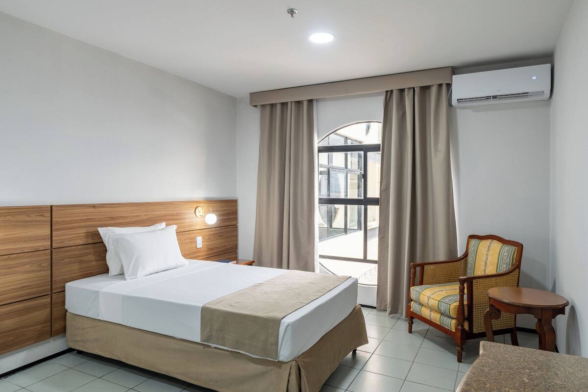 A hotel room with a bed, a wooden headboard, a chair, a small table, and an air conditioning unit.