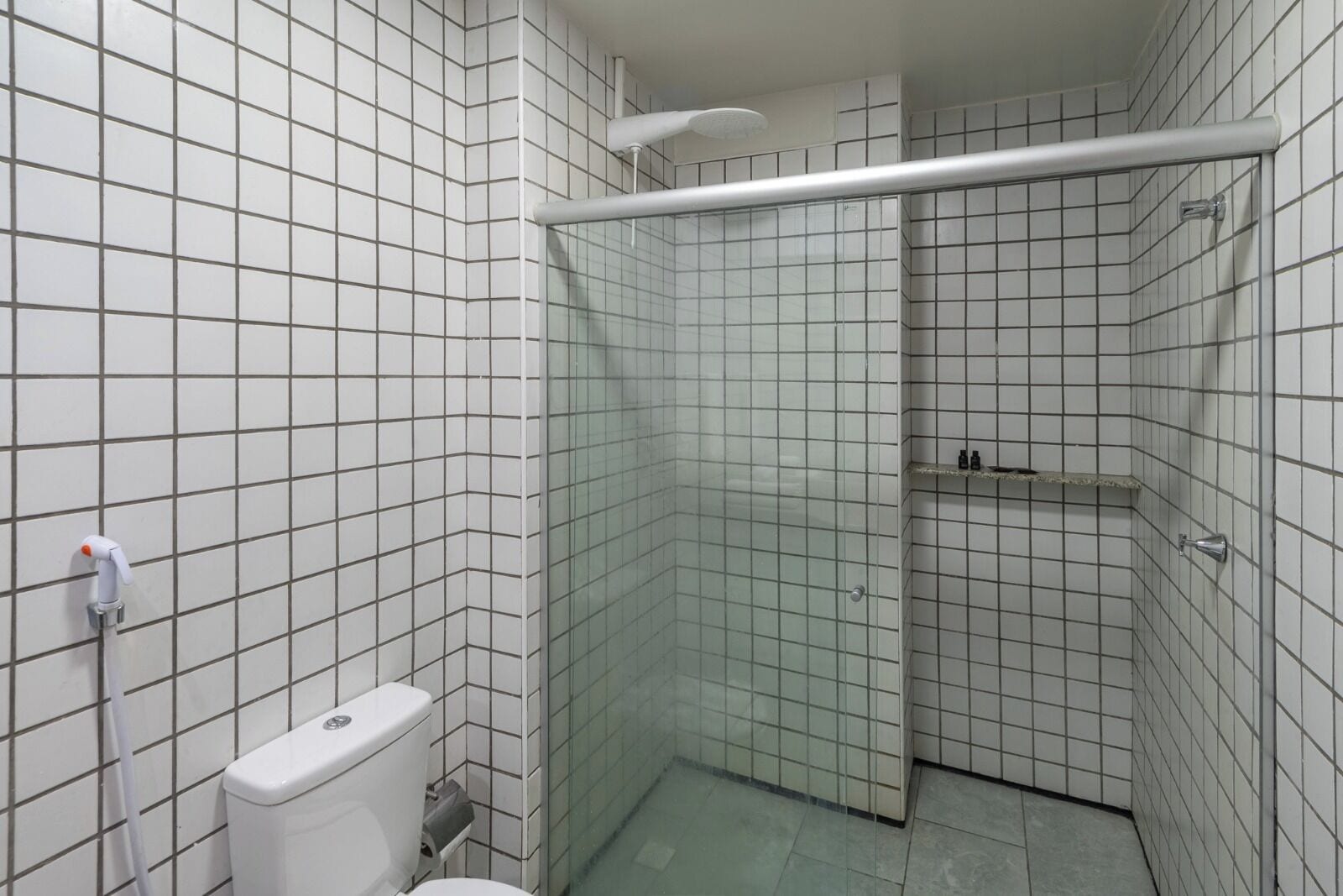 Um banheiro pequeno com paredes e chão em azulejos brancos, vaso sanitário, box de vidro para chuveiro e porta-toalhas de metal.