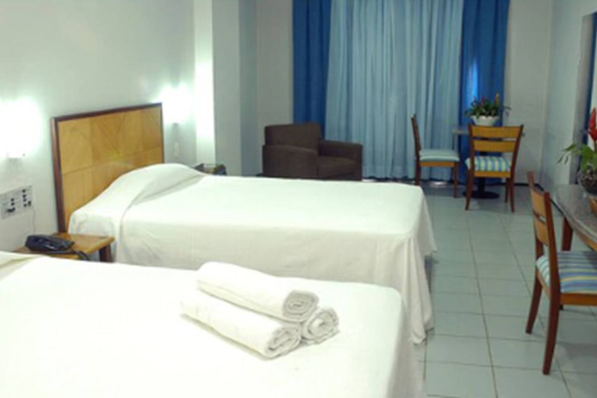 A hotel room with two beds, a desk with a chair, a small table with a potted plant, and a window with blue curtains.
