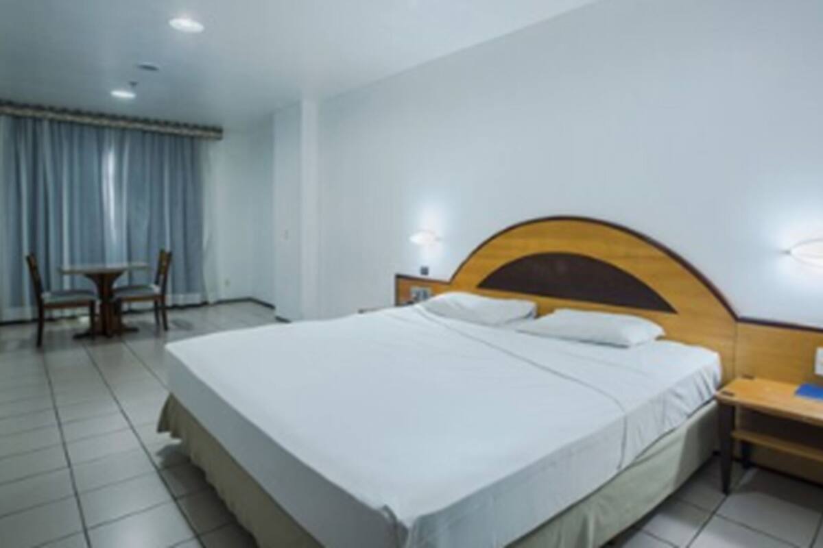 A hotel room with a large bed, a wooden headboard, two bedside tables, a chair, and a window with curtains.