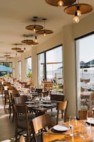 Um restaurante com várias mesas e cadeiras, janelas amplas e luminárias pendentes.