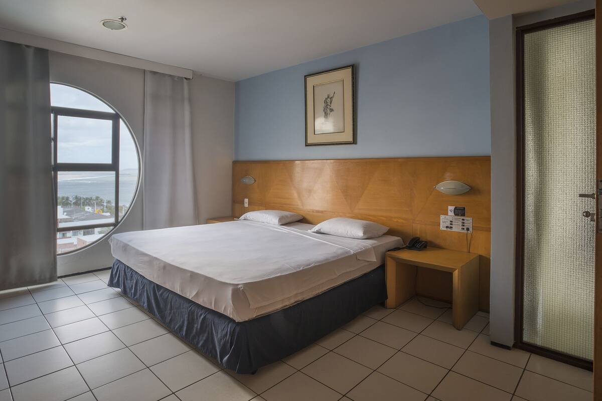 A hotel room with a large bed, a nightstand with a phone, a wall-mounted light, a framed picture, and a view of the ocean through a window.