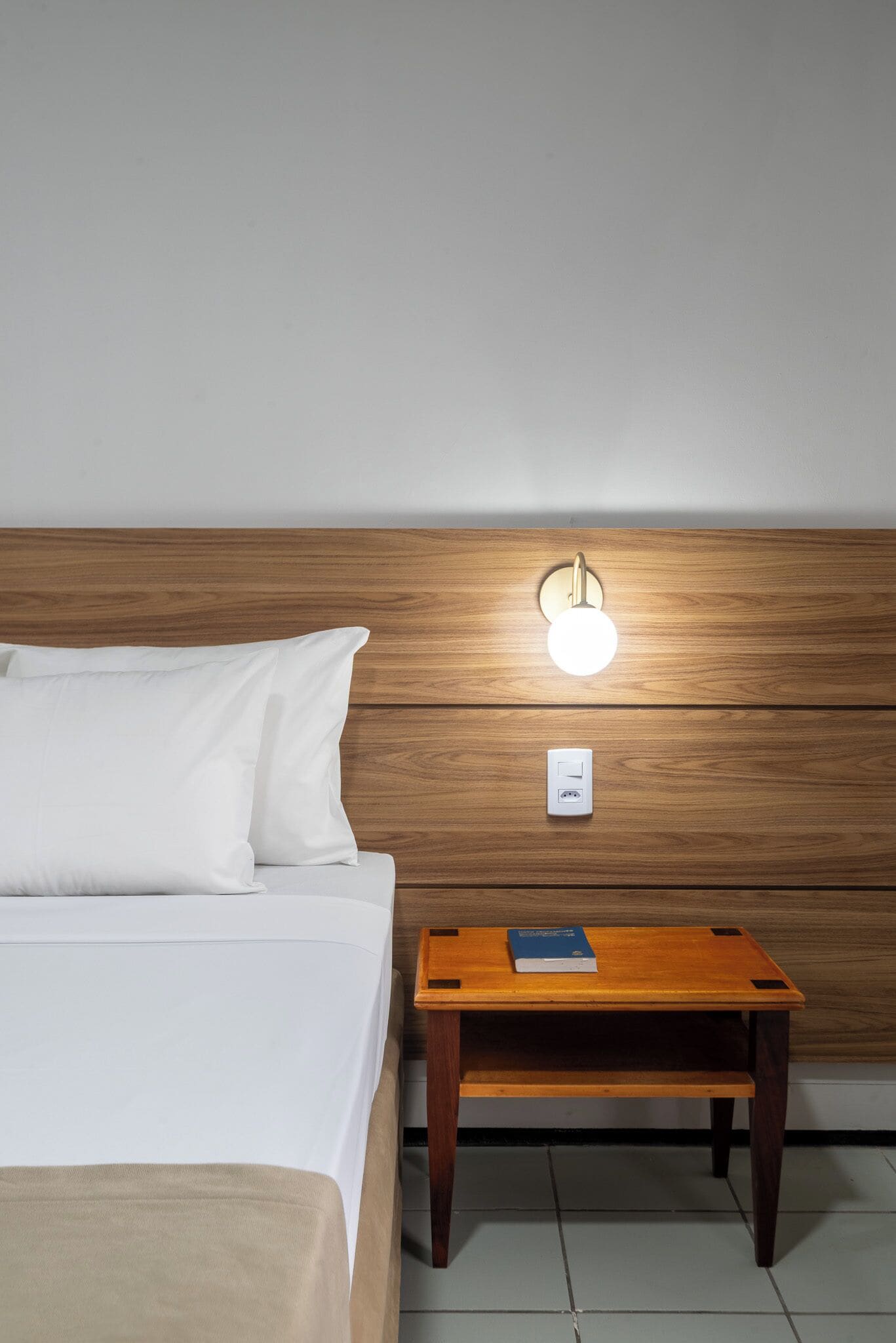 A neatly made bed with white bedding, a wooden nightstand with a book and a wall-mounted light, and a power outlet.