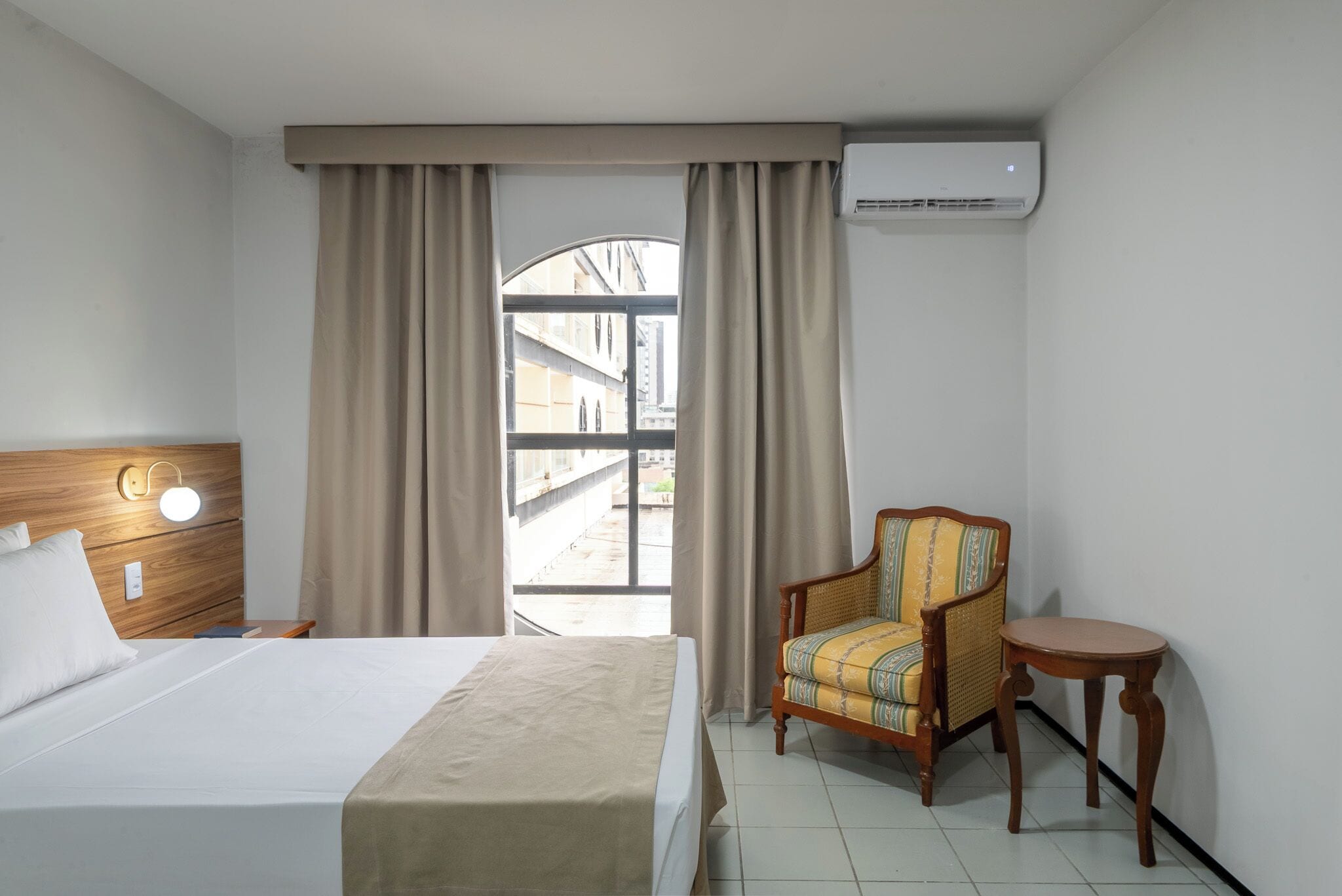 A hotel room with a bed, a chair, a small table, and an air conditioning unit.