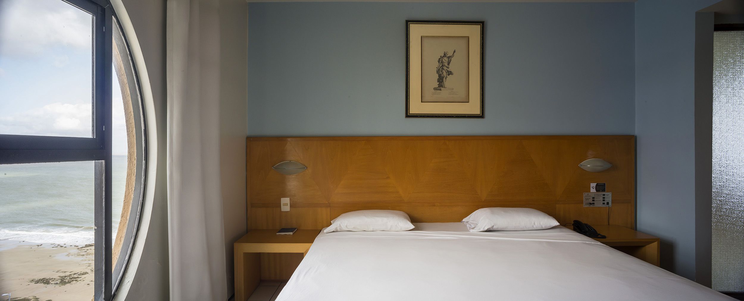 Quarto de hotel com cabeceira de madeira, cama com roupa de cama branca, mesa de cabeceira com abajur, quadro na parede e vista para a praia pela janela.
