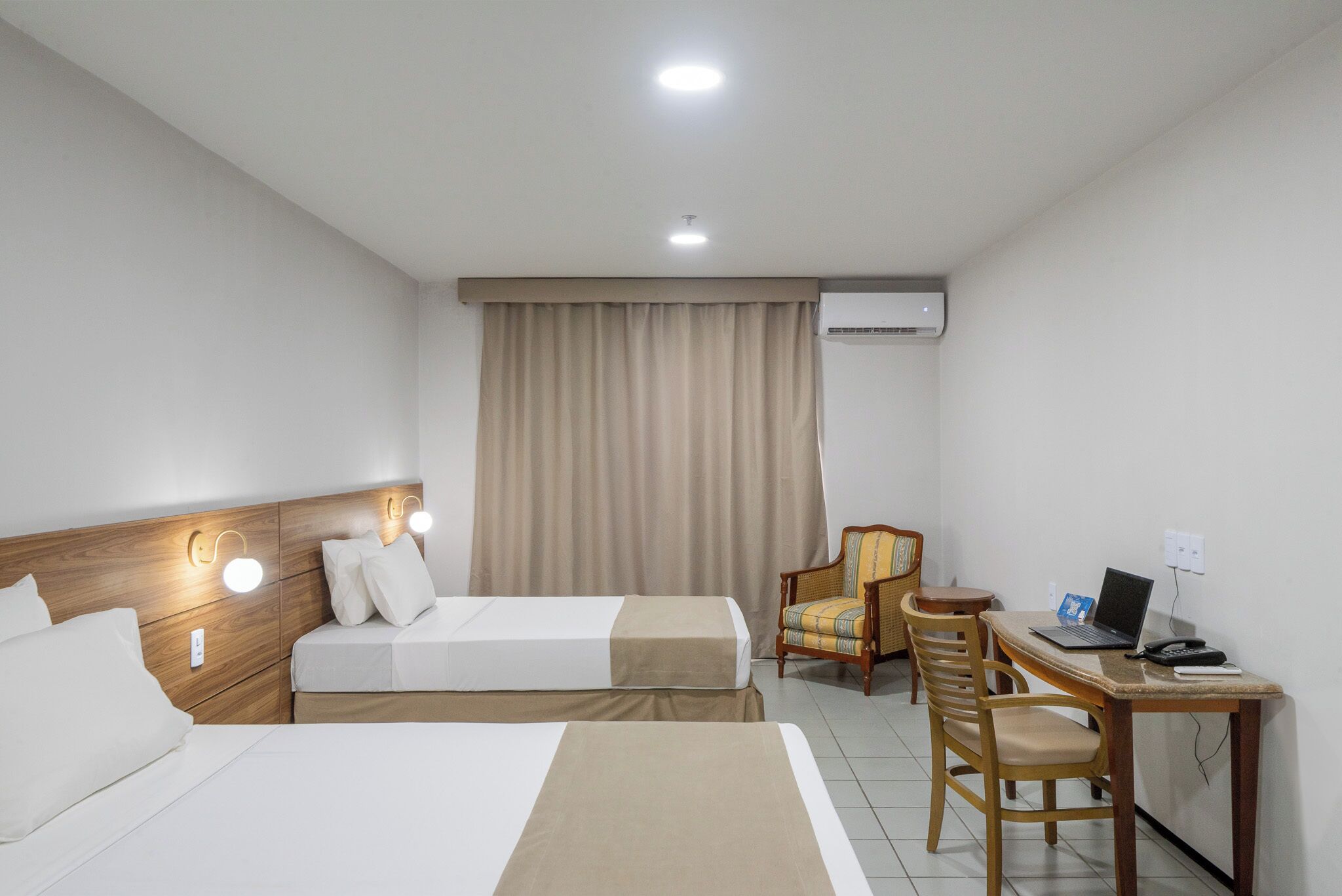 A hotel room with two beds, a desk with a laptop, a chair, and an air conditioning unit.