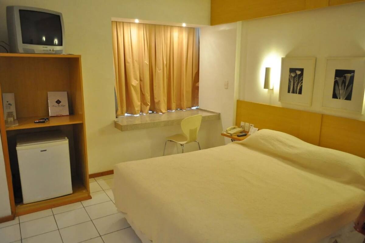 A hotel room with a bed, a desk, a chair, a small refrigerator, and a television on a shelf.