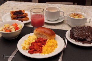 Un desayuno con tostadas, ensalada de frutas, huevos revueltos, salchichas y un pastel de chocolate sobre una mesa de mármol.