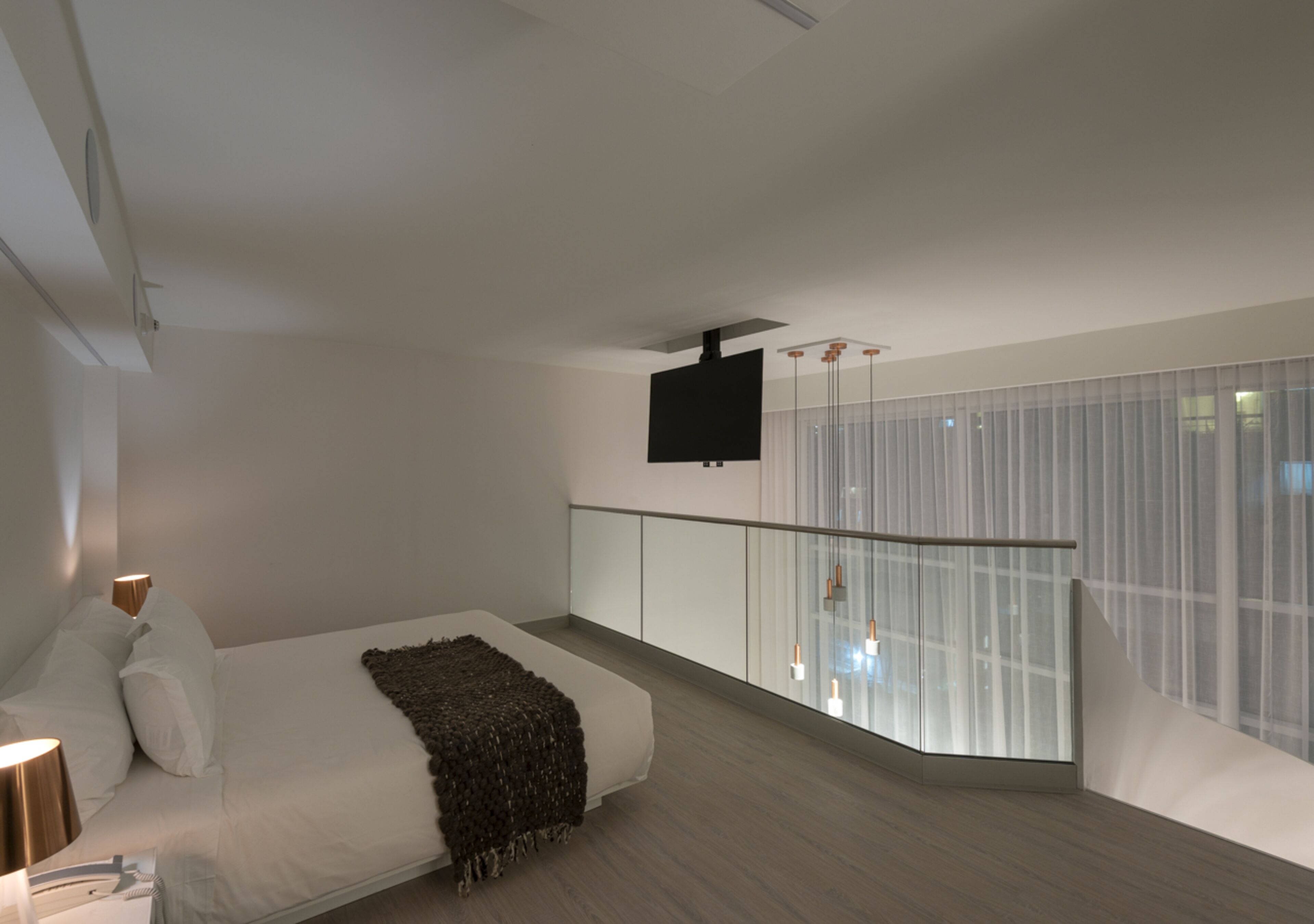A modern bedroom with a large bed, a TV mounted on the wall, and a balcony with a glass railing.