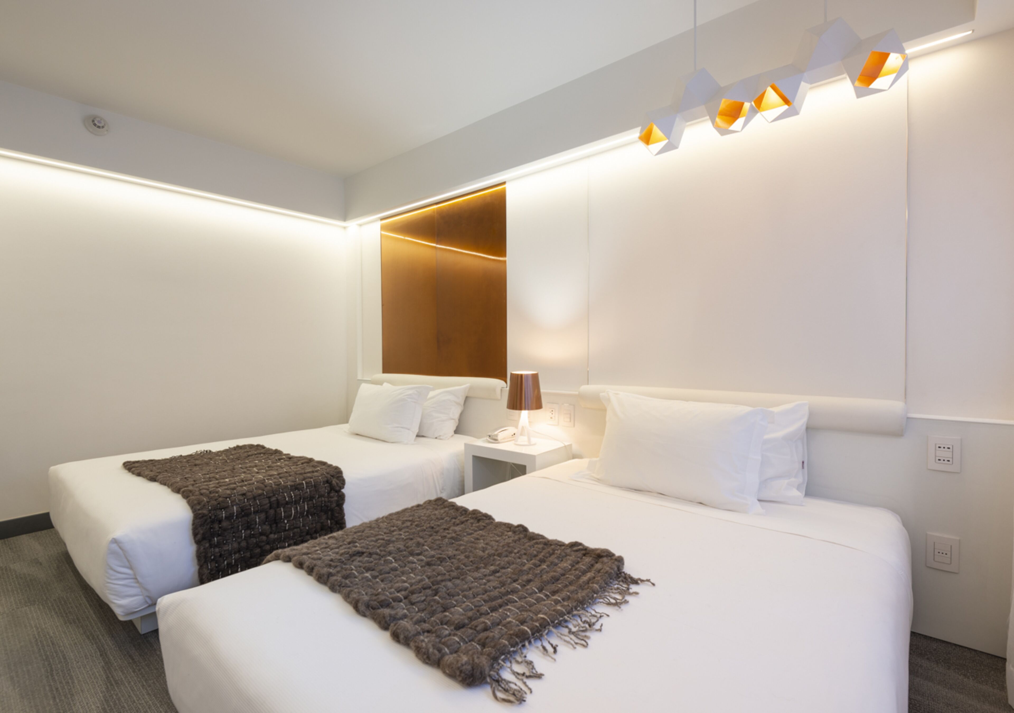 Habitación de hotel con dos camas, una mesita de noche con lámpara y una lámpara moderna sobre las camas.